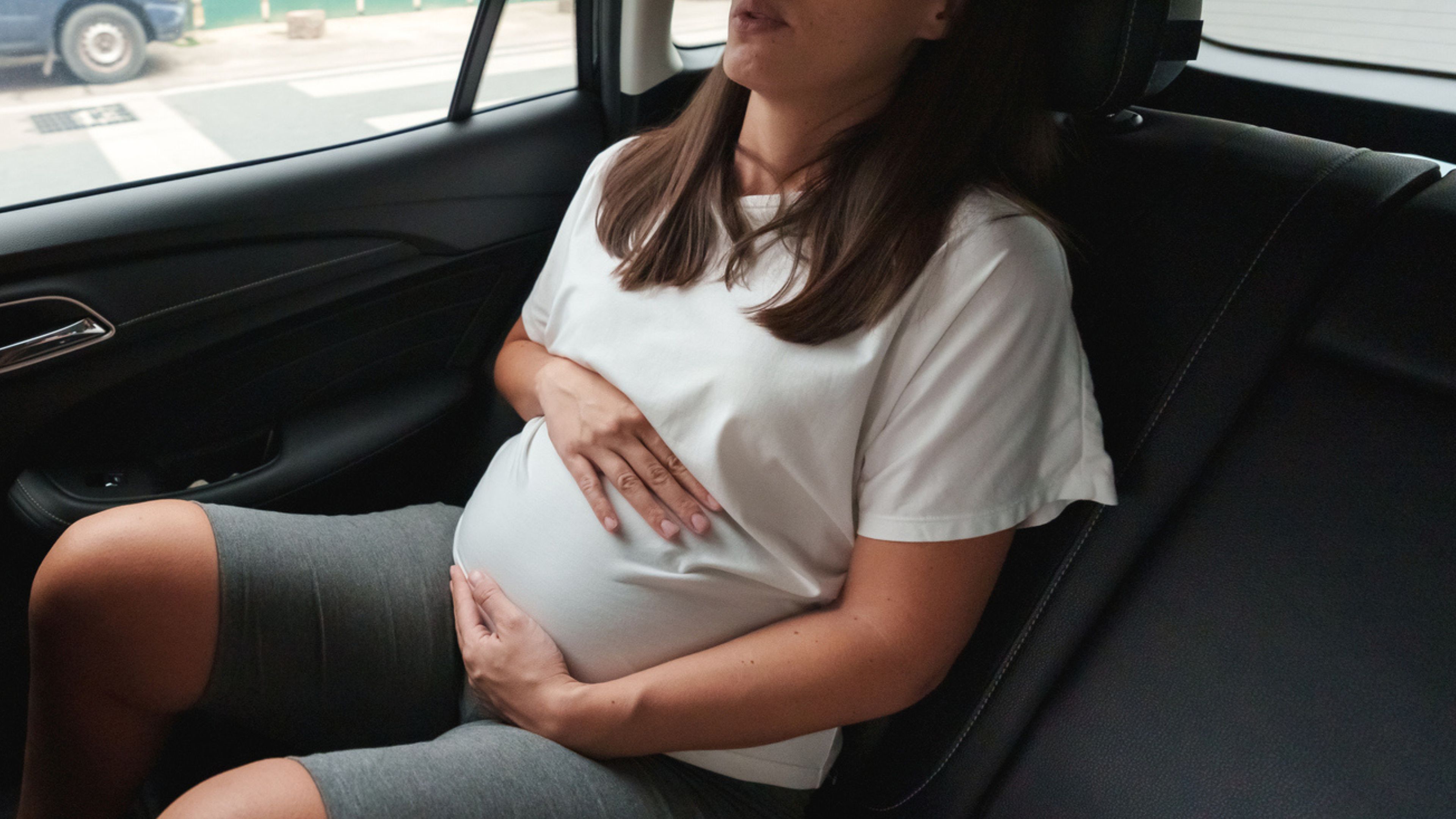 A pregnant woman sitting in the back seat of a car, cradling her stomach.