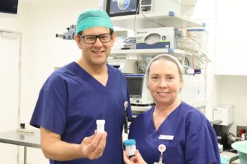 A man and a woman in scrubs in a theatre, holding sample pots.