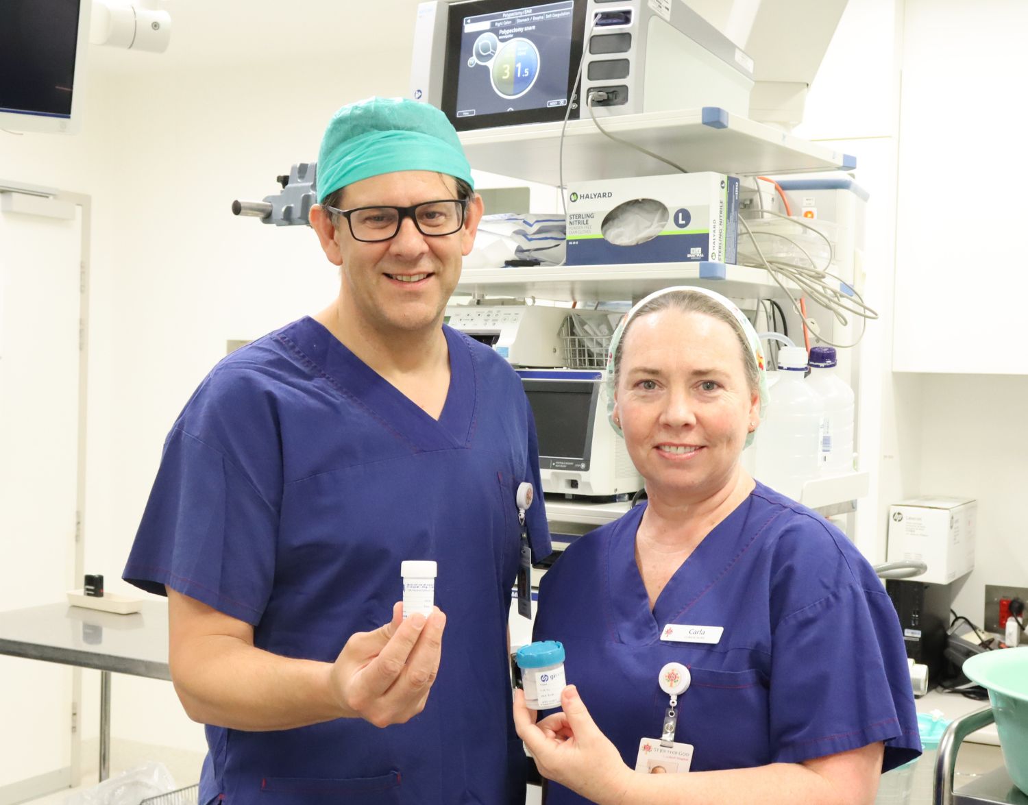 A man and a woman in scrubs in a theatre, holding sample pots.