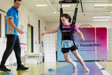 A girl in a harness, suspended from above, watched over by a male support worker.