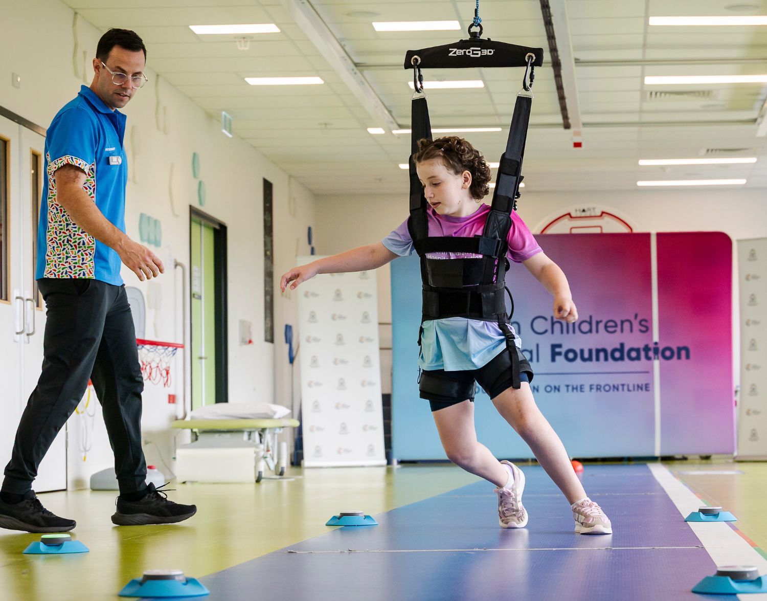 A girl in a harness, suspended from above, watched over by a male support worker.